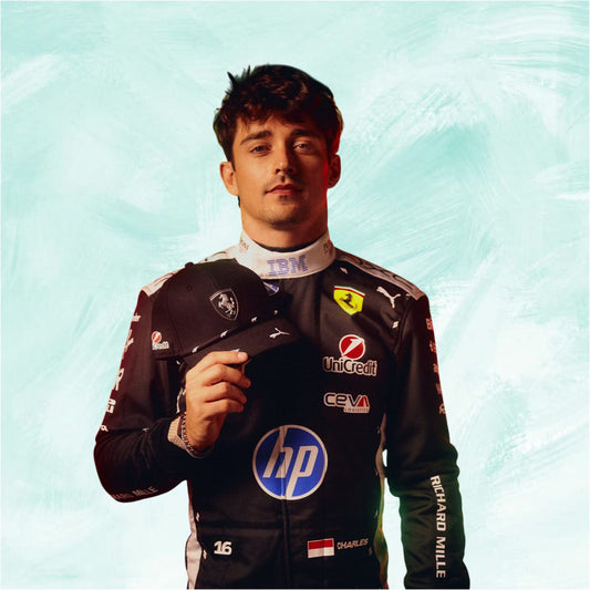 A portrait of Formula 1 driver Charles Leclerc wearing a black Ferrari racing suit with sponsor logos, holding a black cap against a textured light blue-green background.