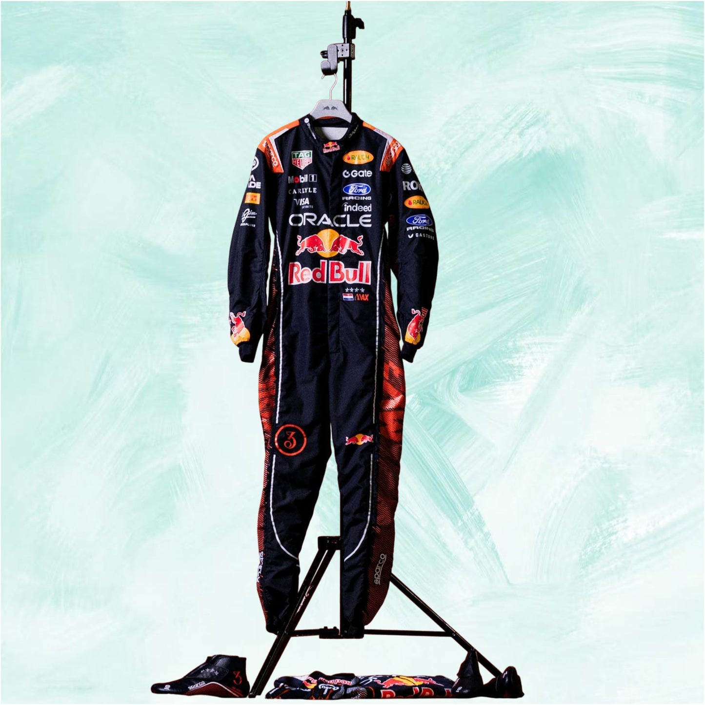 A full Red Bull racing suit displayed on a stand, with matching gloves and a folded suit placed below on the floor. The suit is predominantly black with red and orange graphics and multiple sponsor logos including Oracle, Red Bull, and Castore, set against a light mint green textured background.