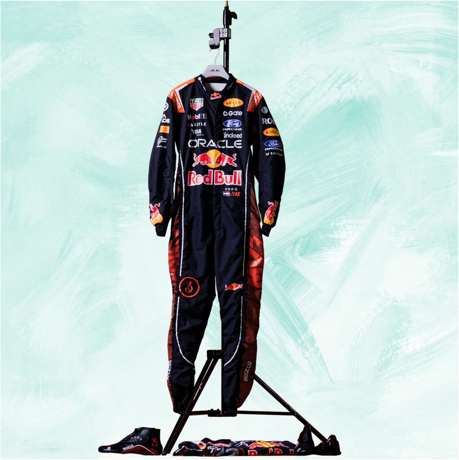 A full Red Bull racing suit displayed on a stand, with matching gloves and a folded suit placed below on the floor. The suit is predominantly black with red and orange graphics and multiple sponsor logos including Oracle, Red Bull, and Castore, set against a light mint green textured background.